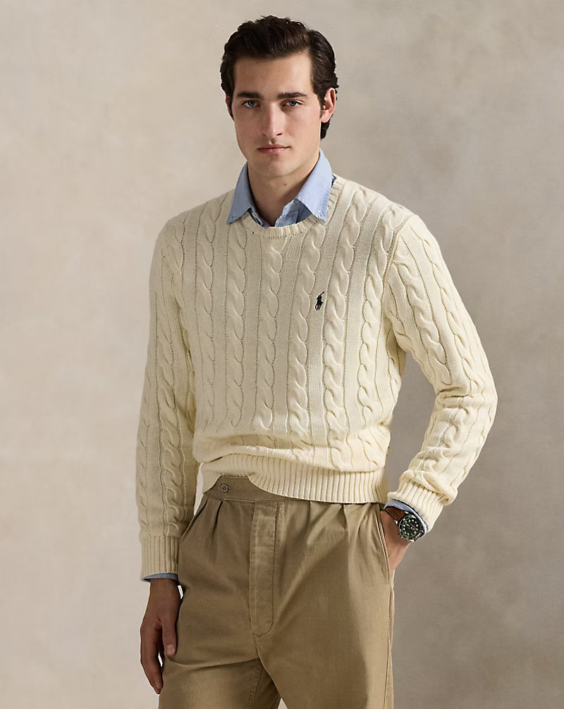 Man wearing a cream cable knit sweater with a brand logo, beige pants, and a light blue shirt underneath.